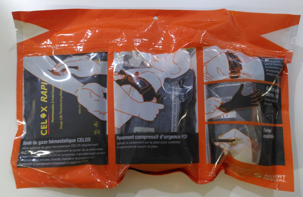 Paquet de plàstic d'un kit de contenció d'hemorràgies francés. D'un color taronja metàl·lic és una bossa de plàstic rectangular segellada a la part superior. Aquest paquet està segmentat en tres espais amb una finestra transparent a cada segment. En el de l'esquerra hi ha un paquet fosc de CELOX RAPID amb una llegenda a sota que diu: "Bande de gaze hémostatique CELOX" i les instruccions de com usar-lo que són il·legibles a la imatge. També hi ha una imatge d'un avantbraç amb una ferida i una altra mà que fica una gasa (la del producte de dins) dins la ferida. En el segment central hi ha una imatge d'un tors i cap humà de perfil amb unes mans que posen un embenat a la part alta del braç, fent referència al contingut del paquet fosc que hi ha dins del segment. També està retolat a sota "Pansement compressif d'urgence FCP" i de nou amb unes instruccions il·legibles en la imatge. En el segment de la dreta hi ha tres dibuixos, un de superior on es veu un braç amb un torniquet aplicat i "Tourniquet" retolat a la dreta. En el centre del segment hi ha un dibuix d'unes mans posant-se un guant i "Gants Nitrile, Large" retolat i a la part baixa del segment hi ha dibuixada una mà sostenint un retolador petit i retolat com a "Feutre indélébile".