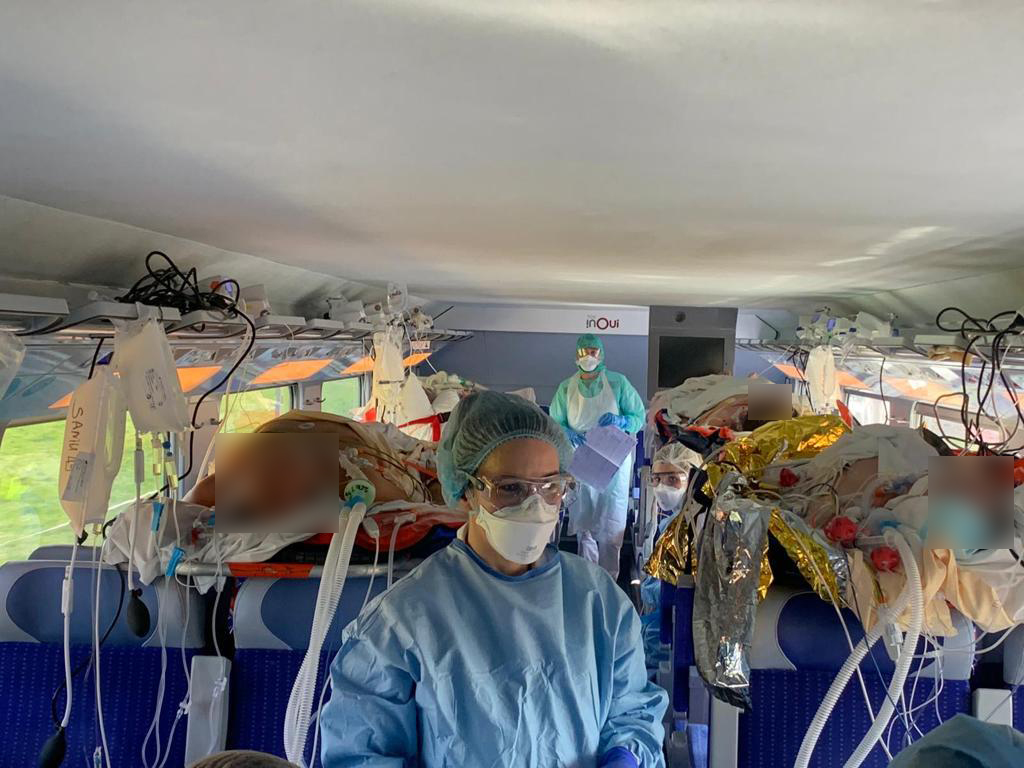 Interior d'un tren TGC medicalitzat per al trasllat de pacients COVID. En primer pla, en el passadís entre seients d'un vagó, es veu una professional sanitària amb l'EPI complert (mascara FFP, casquet, ulleres, bata i guants). A ambdos costats i en lliteres sobre els seients hi ha diversos pacients intubats. Molt equipament mèdic es troba als suports per deixar les malestes. Al fons de la imatge s'hi veu un altre professional sanitari.