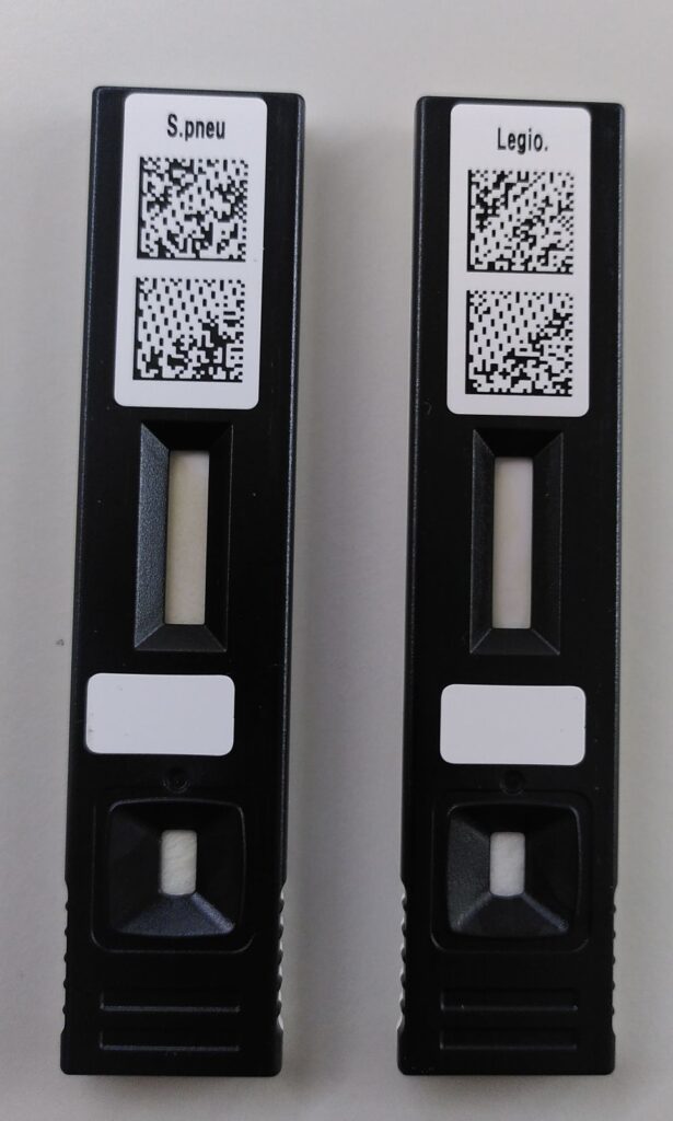 Cartutx d'un parell de tests d'antígens. Dues capsetes de plàstic negre allargades, a la part superior tenen una etiqueta amb les abreviatures S.pneu i Legio i uns codis QR a sota. En el centre del cartutx hi ha el rectangle de lectura de resultats, complemaent blancs els dos. Una mica per sota hi ha el quadradet per a posar-hi les gotes de la mostra